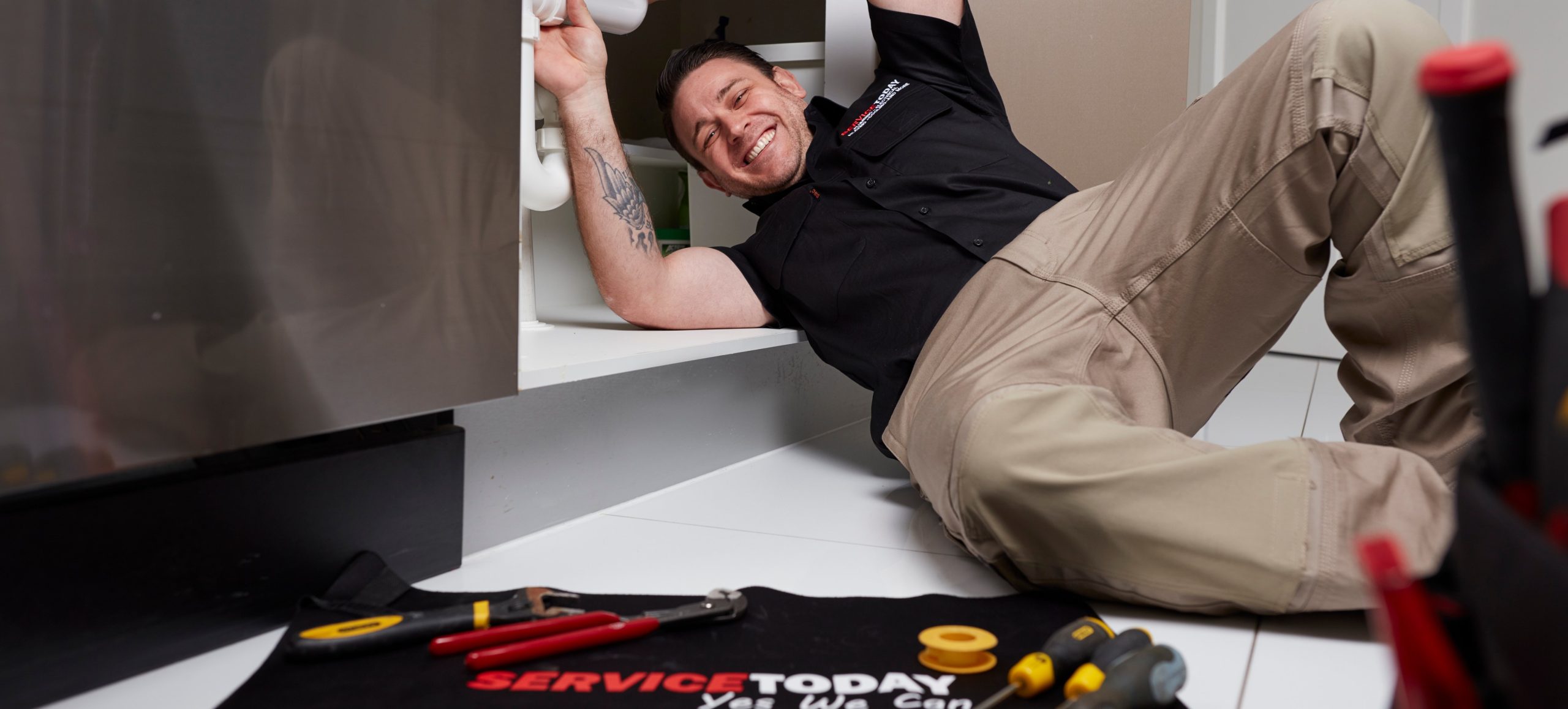 Plumber working under a sink, smiling, with tools on a mat.